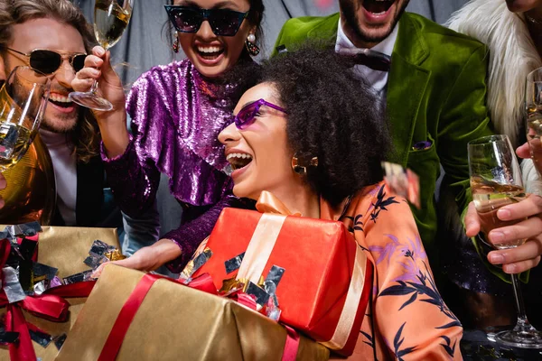 笑容满面的非洲裔美国女人 戴着礼品盒 坐在灰色背景的跨种族朋友中间 — 图库照片