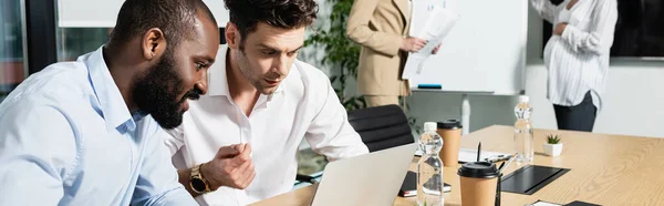 interracial businessmen looking at laptop near businesswomen working during meeting, banner