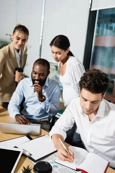 Hombre Negocios Escribiendo Cuaderno Cerca Colegas Interracial Trabajando Sobre Fondo —  Fotos de Stock