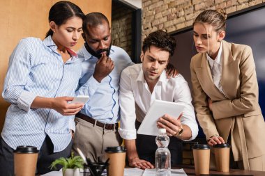 serious businessman holding digital tablet near thoughtful colleagues during meeting clipart