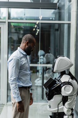 Afro-Amerikan mühendis ofisteki insansı robota bakıyor.