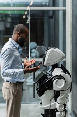 Gülümseyen Afrikalı Amerikalı iş adamı ofiste robotun yanında el kol hareketi yapıyor.