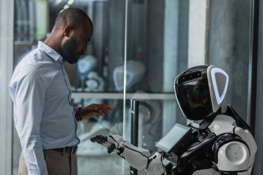 Afrika kökenli Amerikalı iş adamı ofiste insansı robotun yanında duruyor.