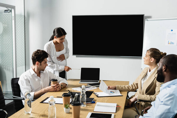 беременная американская бизнесвумен указывает на ноутбук рядом с коллегами