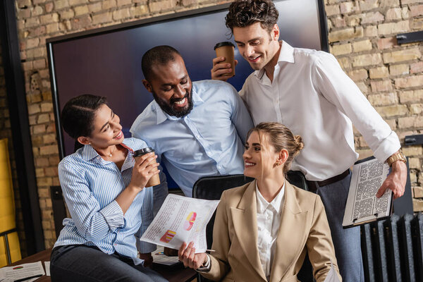 happy businesswoman holding newspaper near smiling multiethnic colleagues