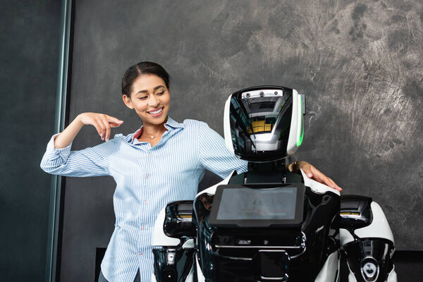 joyful african american businesswoman pointing at humanoid robot in office