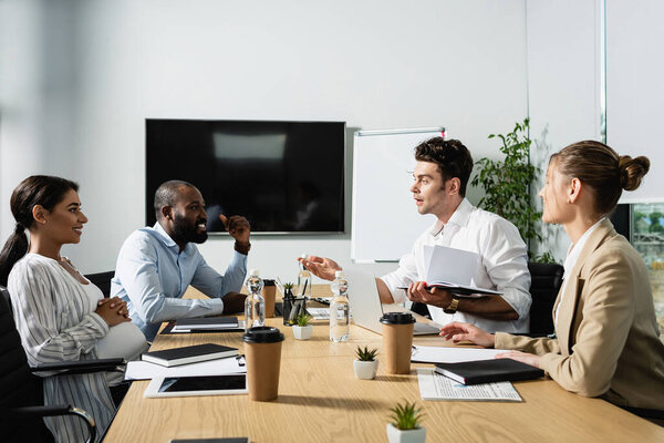 businessman pointing with hand while talking to smiling multicultural colleagues