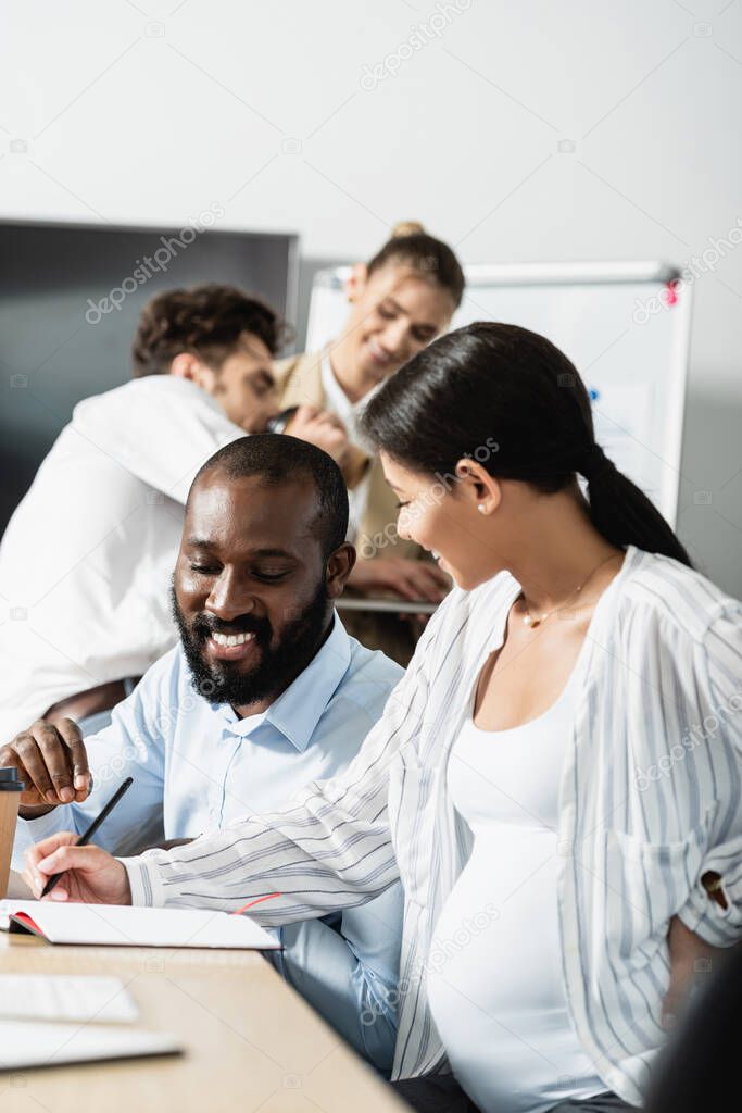 pregnant businesswoman writing in notebook near african american colleague and blurred coworkers