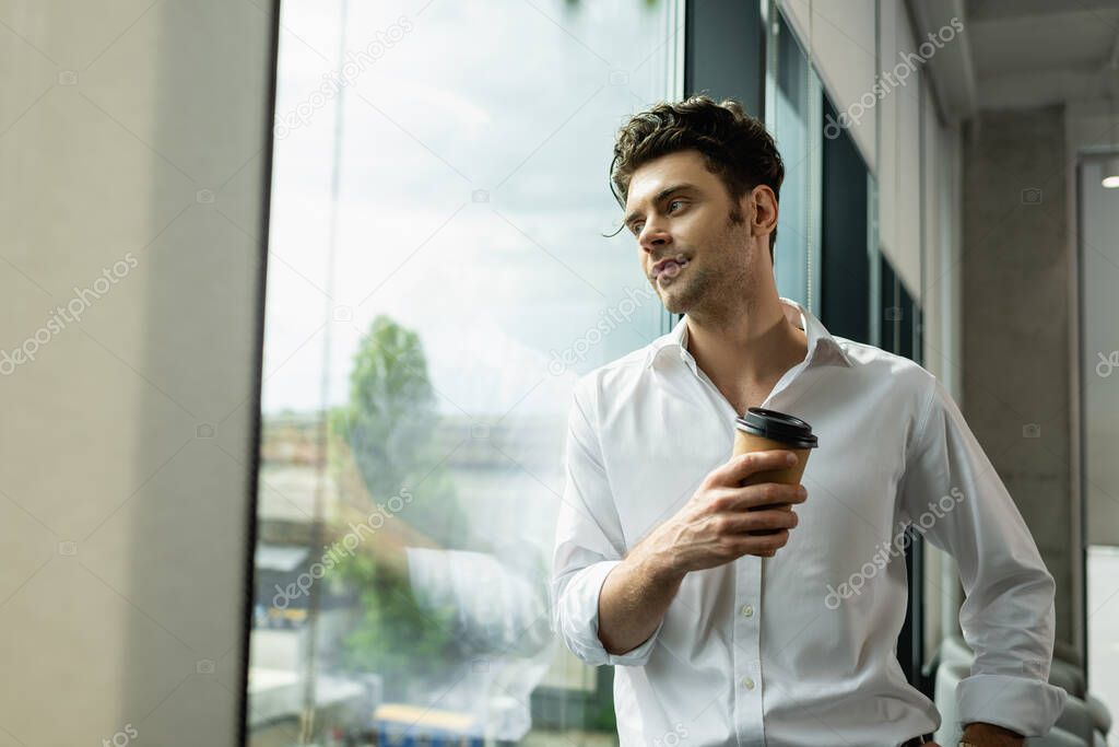 positive, dreamy businessman standing near window with coffee to go