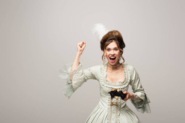 КИЕВ, УКРАИНА - 22 апреля 2021 года: взволнованная женщина в винтажном платье показывает жест победы, держа геймпад изолированным на сером