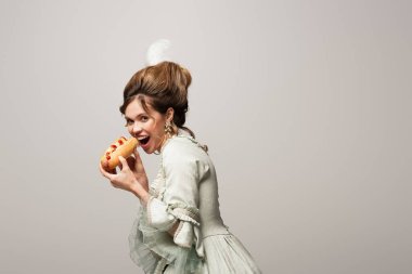Gri renkte izole edilmiş lezzetli sosisli yiyen eski moda kıyafetli aç bir kadın.