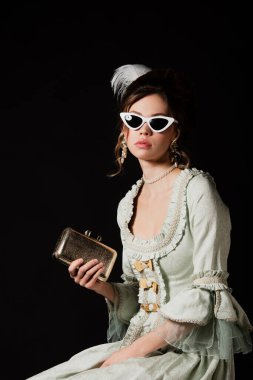 elegant woman in vintage dress and trendy sunglasses posing with golden purse bag isolated on black clipart
