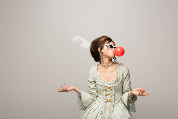 stylish woman in retro dress and trendy sunglasses blowing bubble gum isolated on grey
