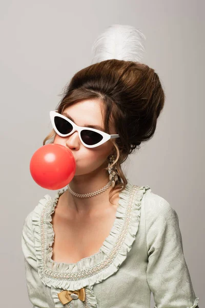 vintage style woman in trendy sunglasses blowing red bubble gum isolated on grey
