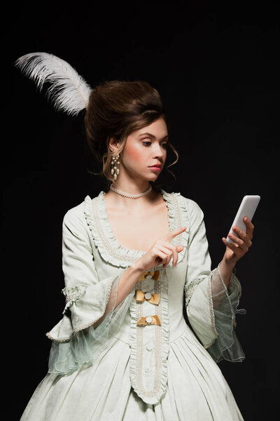 pretty woman in vintage outfit pointing at mobile phone isolated on black