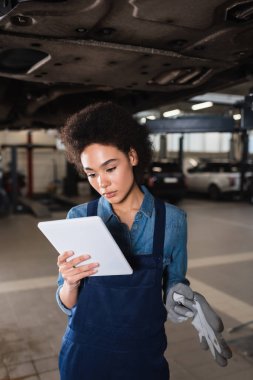 Genç Afro-Amerikan tamirci arabanın altında duruyor ve garajda dijital tablet tutuyor.