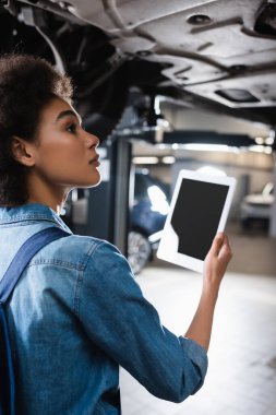 Ciddi bir Afro-Amerikalı tamirci arabanın altında dikiliyor ve elinde garajda boş ekranlı dijital tablet tutuyor.