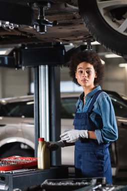 Genç Afro-Amerikan tamirci arabanın altında duruyor, elinde motor yağı şişesiyle garajdaki kameraya bakıyor.