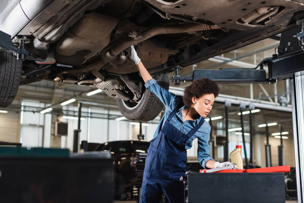 young african american mechanic in overalls repairing bottom of car in garage