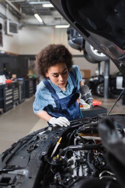 Genç Afro-Amerikan tamirci elinde elektrikli tornavidayla garajda açık kaputlu bir arabaya bakıyor.