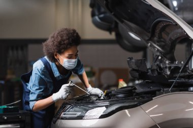 Korumalı maskeli genç Afro-Amerikan tamirci garajda arabanın motoruyla çalışıyor.