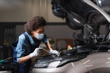 young african american mechanic in protective mask working with motor of car in garage clipart