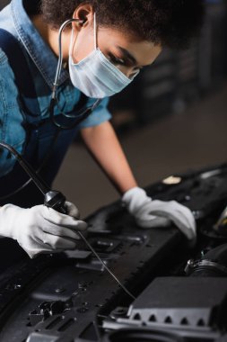 Koruyucu maskeli, eldivenli ve steteskoplu genç Afro-Amerikan tamirci garajda araba motoruyla çalışıyor.