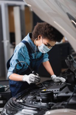 Koruyucu maskeli ve steteskoplu genç Afro-Amerikan tamirci garajda araba motoruyla çalışıyor.