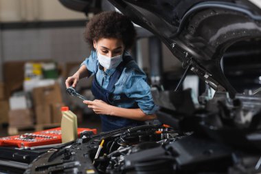 young african american mechanic in protective mask working with car motor in garage clipart