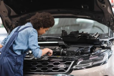 Genç Afro-Amerikan tamirci garajda kaputu açık araba motoruyla çalışıyor.