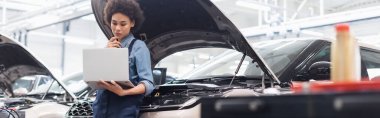 serious young african american mechanic holding laptop near car with open hood in auto repair service, banner clipart