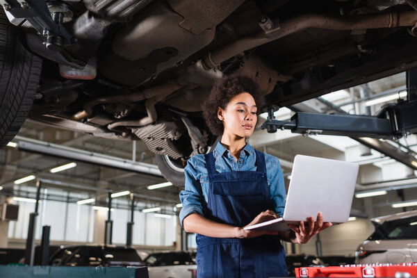 young african american mechanic standing underneath car and holding laptop in garage