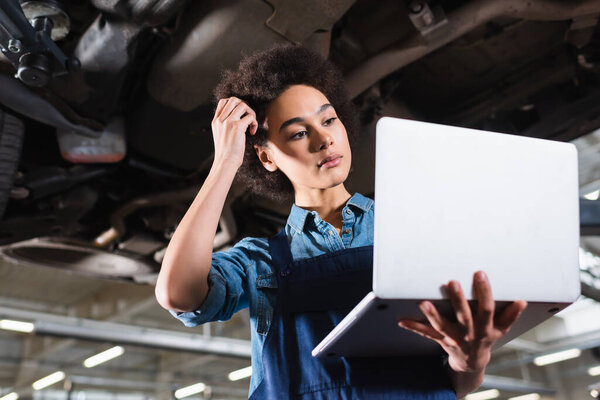 young african american mechanic standing underneath car with laptop and holding hand near head in garage