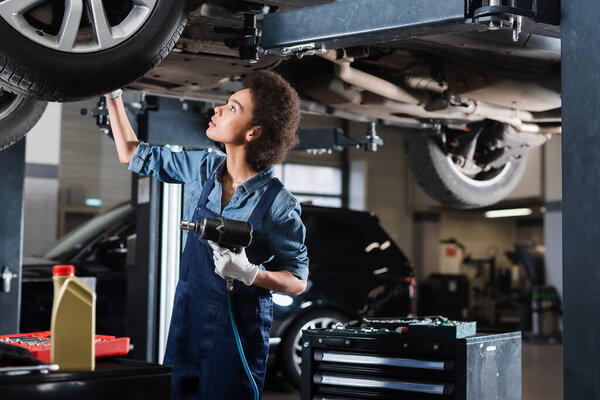 young african american mechanic in overalls repairing lifted car with electric screwdriver in garage