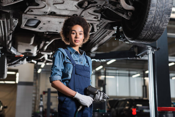 young african american mechanic in overalls standing with electric screwdriver underneath lifted car in garage