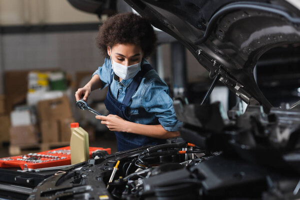 young african american mechanic in protective mask working with car motor in garage