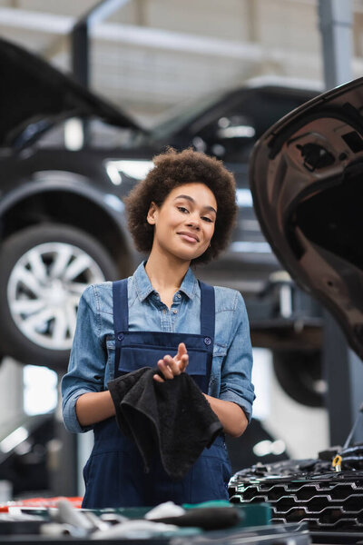 smiling young african american mechanic in overalls drying hands with black towel in garage