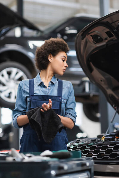 young african american mechanic in overalls drying hands with towel in garage