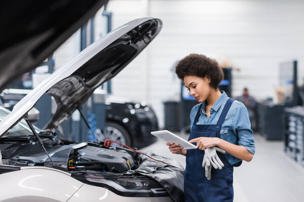 young african american mechanic holding digital tablet near car with open hood in auto repair service