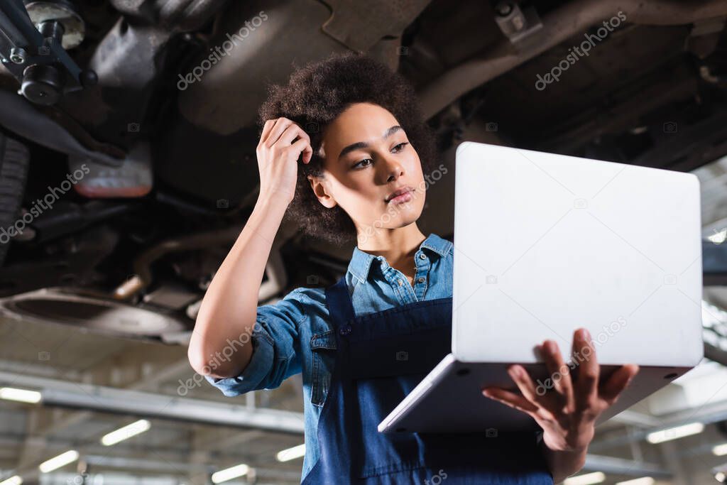 young african american mechanic standing underneath car with laptop and holding hand near head in garage