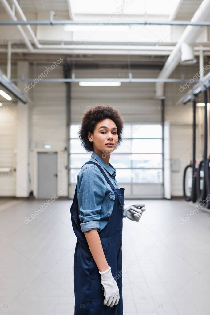 confident young african american mechanic in gloves standing and looking at camera in garage