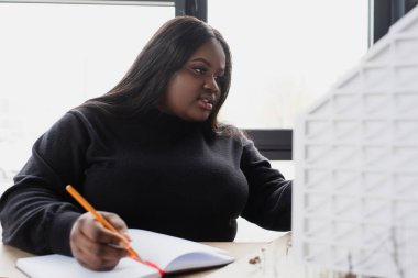 Afro-Amerikan artı boyut tasarımcısı ev modeline bakarken deftere yazıyor. 