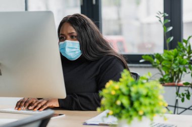 Afro-Amerikan artı tıbbi maskeli iş kadını bilgisayar monitörünün yanında klavyede yazı yazıyor.