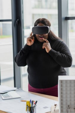 VR kulaklıklı Afro-Amerikan kadın şaşırdı. Masa başındaki karton ev modeli ve taslağı.