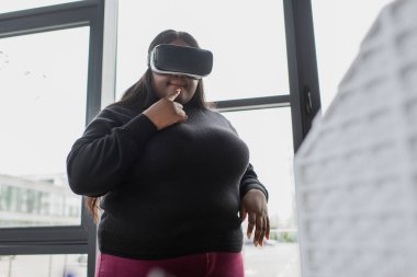 Kafası karışık Afrikalı Amerikalı artı boyut mühendisi vr kulaklık, bulanık ev modelinin yanında. 