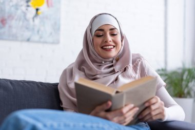 Neşeli Müslüman kadın evde kitap okuyor, ön planı bulanık.
