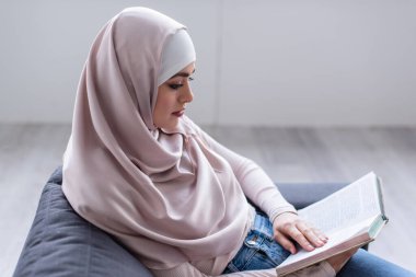 Evde tesettürlü genç Arap kadın kitap okuyor.