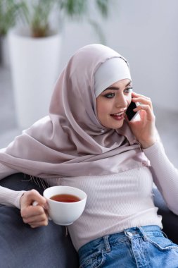 Kafiyeli genç Arap kadın elinde çay fincanı tutarken akıllı telefondan konuşuyor.