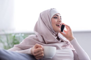 Bir fincan çayla evde akıllı telefondan konuşan neşeli Müslüman kadın.