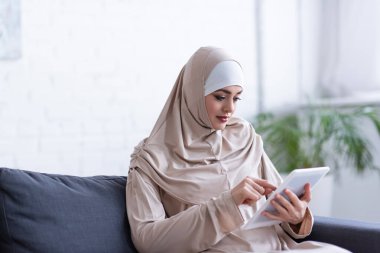 Genç Müslüman kadın oturma odasındaki kanepede dijital tablet kullanıyor.